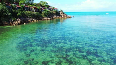 Beautiful-pattern-of-seabed-with-coral-reefs-growing-under-crystal-emerald-water-washing-rocky-shoreline-of-tropical-island-with-beach-houses-in-Thailand