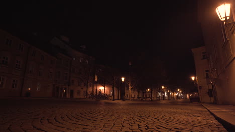Empty-city-square-Kampa-of-paving-stones-in-Prague,-Czechia,-with-trees,-old-houses-and-street-lanterns-shining-above-during-a-Covid-19-lockdown-at-night,-illuminated-by-streetlights,-slow-pan-shot