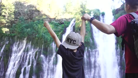 Vater-Und-Sohn-Heben-Gemeinsam-Die-Hände,-Während-Sie-Händchen-Haltend-Vor-Einem-Wunderschönen-Wasserfall-Stehen