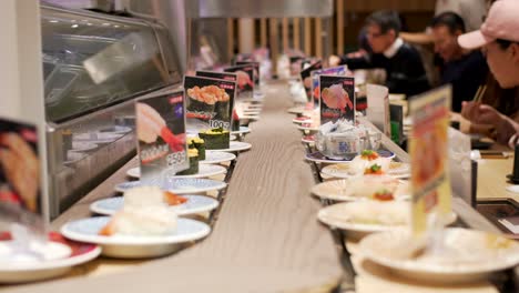 Platos-De-Sushi-En-La-Cinta-Transportadora-Con-Clientes-Recogiendo-Y-Comiendo-Dentro-De-Un-Restaurante-Kaitenzushi---Sushi-De-Cinta-Transportadora-En-Kyoto,-Japón