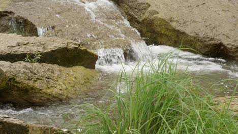 Clear-shallow-fresh-water-stream-flows-over-rocks-on-sunny-day,-close-up-static-slow-motion