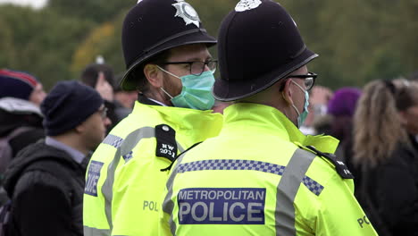 Two-police-officers-in-protective-face-masks,-both-wearing-glasses,-stand-monitoring-a-crowd-of-people-attending-a-Coronavirus-and-QAnon-conspiracy-protest-and-ignoring-social-distancing-restrictions