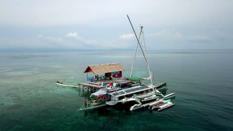 Bigkanu-Trimaran-Segelboot-An-Einem-Ferienhaus-Am-Meer-Mit-Mann,-Der-Vom-Schiff-Aus-Ins-Wasser-Taucht,-Luftbild-Um-Den-Kreis-Herum