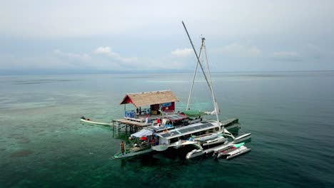 Velero-Trimarán-Bigkanu-En-Una-Cabaña-Oceánica-Con-Un-Hombre-Buceando-En-Las-Aguas-Desde-El-Barco,-Círculo-Aéreo-Alrededor-Del-Tiro