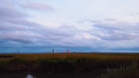 HD-MOTIONLAPSE-Zoom-in-and-hold-Atlantic-City-skyline-in-distance-over-waterway-with-mostly-cloudy-sky-day-to-night-with-clouds-turning-pink,-purple-and-blue