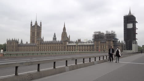 Two-people,-one-wearing-a-surgical-face-mask,-walk-across-a-near-deserted-Westminster-Bridge-and-past-the-house-of-Parliament-during-the-Coronavirus-outbreak