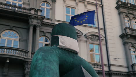 Sculpture-made-of-metal-symbolizing-freedom-with-a-veil-guise-over-mouth,-symbolizing-quarantine-and-protection-against-the-Coronavirus-disease-with-a-waving-Europe-Union-flag-from-captured-in-120fps