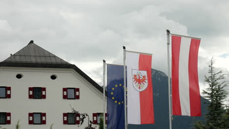 Flaggen-Europas,-Tirols-Und-Österreichs-Wehen-Im-Wind,-Mit-Hotel-Im-Hintergrund-An-Bewölkten-Tagen