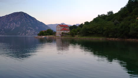 A-luxurious-villa-with-a-small-pier-and-a-garden-on-the-shores-of-Kotor-bay-in-Montenegro-surrounded-by-trees,-small-coastal-town-behind,-mountains-on-the-horizon-around-the-bay,aerial-fly-above-shot