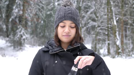 Young-caucasian-woman-drinking-hot-tea-from-thermo-bottle-in-the-park-on-snowy-day