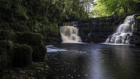 Time-lapse-of-dark-spring-forest-cascade-waterfall-surrounded-by-trees-with-rocks-in-the-foreground-in-rural-landscape-of-Ireland