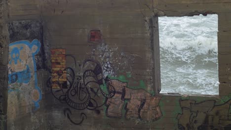 View-to-the-stormy-sea-through-the-window-of-abandoned-coast-defense-building-with-graffiti