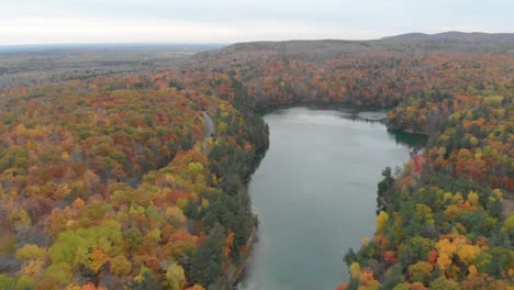 Umkehrende-Luftaufnahmen-über-Dem-Pink-Lake-In-Gatineau,-Quebec,-Mit-Bergen-Und-Dem-Blauen-Wasser-In-Der-Ferne-Sowie-Aussichtspunkten-Und-Hügeln-Mit-Herbstlichen-Waldbäumen-Auf-Beiden-Seiten