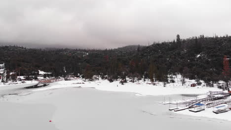 Aerial-drone-flyover-of-Frozen-Lake-on-overcast-winter-day,-heading-towards-forested-mountain-range