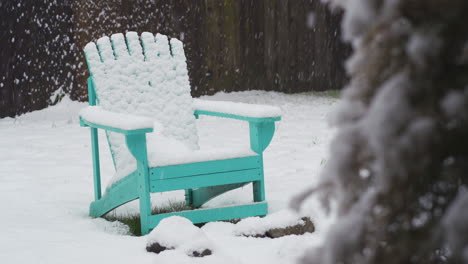 Still-shot-of-snow-falling-in-snowy-backyard-on-teal-lawn-chair-in-front-of-wooden-fence