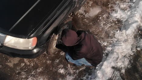 Man-Kneeling-Down-to-Check-Tire-in-Snowy-Driveway-AERIAL