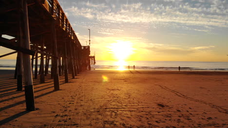 Drohne-Fliegt-Bei-Sonnenuntergang-Unter-Dem-Newport-Beach-Pier,-Während-Das-Sonnenlicht-Strahlt-Und-Menschen-In-Silhouetten-Luftbildern-An-Der-Kalifornischen-Küste-Spazieren