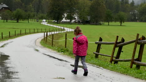 Little-girl-playfully-walking-in-rain-on-country-road,-farmhouse-in-background,-alpine-valley,-steady-shot,-HD,-from-behind-facing-away