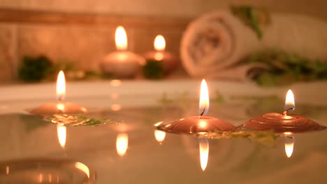 Entspannender-Spa-hintergrund-Mit-Im-Badewasser-Schwimmenden-Kerzen,-Einigen-Grünen-Blütenblättern-Und-Einem-Handtuch-In-Der-Nähe-Der-Wasseroberfläche
