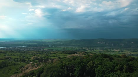 Aerial-drone-shot-of-storm-clouds-over-hills-in-a-valley-with-a-river,-tilting-panomara-view