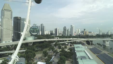 Flyinge-close-by-big-gigantic-ferris-wheel-with-people-on---in-it,-showing-big-city-skyline-of-metropolis-singapore-with-car-traffic,-bridge-,-skysrapers-towers
