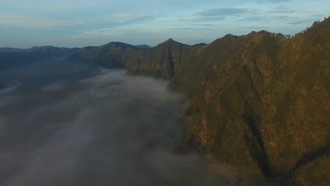 Drone-shot-of-the-Bromo-mountains,-with-a-sea-of-clouds-at-the-bottom-during-sunrise