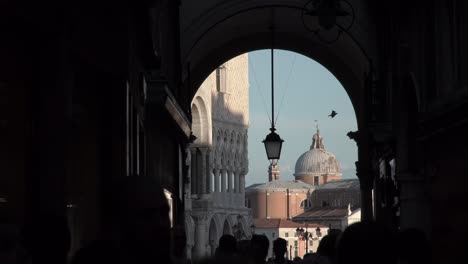 Looking-through-a-arch-creating-a-shadow-with-tourists,-famous-Venice-landmarks-in-the-background