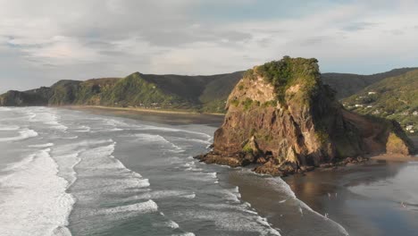Breathtaking-aerial-drone-shot-of-Lion-Rock-on-Piha-Beach-with-surfing-waves-during-sunset