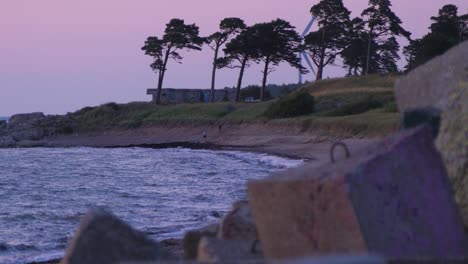 Distant-people-enjoying-romantic-sunset-by-the-sea-with-a-wind-turbine-in-the-background,-wide-shot