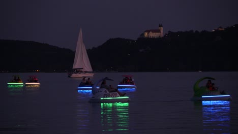 People-on-luminous-paddle-boats,-sailboat-and-a-motorboat-passing-by-at-the-bay-of-Balatonfüred-with-Tihany-Abbey-on-top-of-the-hill-in-the-background-at-dusk
