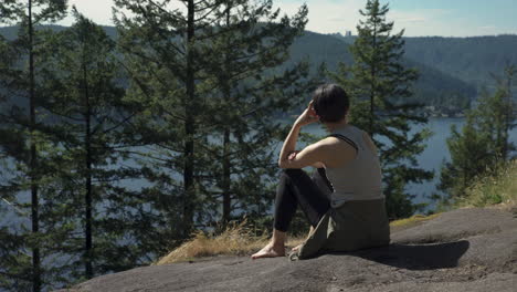 Woman-contemplatively-sitting-on-a-bluff-overlooking-trees,-mountains-and-the-water