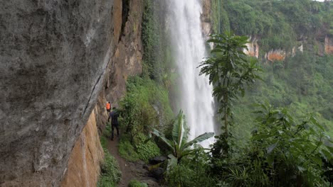Ugandische-Männer-Wandern-Auf-Einem-Schlammigen-Pfad-Hinter-Einem-Großen-Wasserfall-Im-Tropischen-Ostafrika