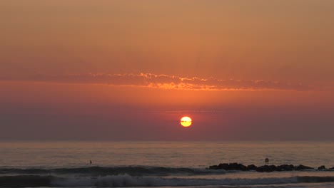Orange-dawn-sunrise-over-calm-sea-with-waves-breaking-on-rocks,-wide-shot