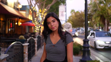 A-young-hispanic-woman-walking-a-downtown-urban-city-street-in-California-with-restaurants,-shops-and-retail-stores-with-lights-at-twilight-SLOW-MOTION