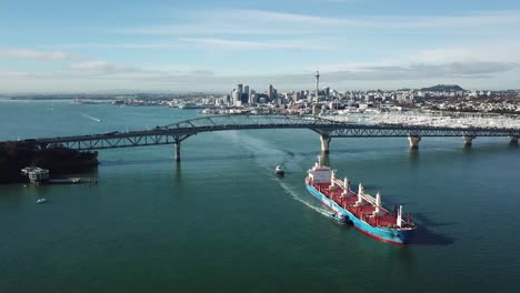 Aerial-view-of-container-cargo-ship-being-guided-by-tug-boats-under-Auckland-harbor-bridge,-New-Zealand-4k