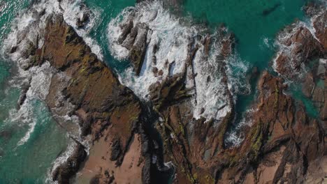 Waves-hit-the-brown-orange-rocks-of-the-shore-with-their-clear-turquoise-water-turning-to-white-foam