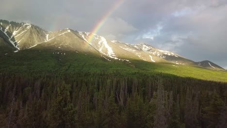 Sensational-Yukon-flight-toward-double-rainbow-arc-by-picturesque-mountain-range-above-green-lush-vegetation-and-forest-trees-on-cloudy-day,-Canada,-overhead-aerial-approach
