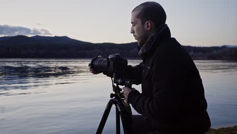 Caucasian-man-adds-filter-to-camera-lens-attached-to-tripod-by-calm-lake-waters-with-dark-mountain-range-in-background,-static-profile-close-up