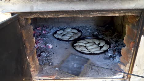 Man-putting-burning-ashes-inside-a-clay-oven-with-a-shovel-near-trays-with-empanadas