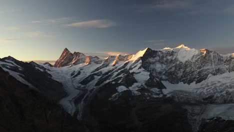 Drone-shot-flying-sideways-to-the-left,-revealing-a-big-mountain-peak-full-of-ice-and-snow-in-the-foreground-of-the-vast-and-bare-landscape-on-top-of-the-alps-in-Switzerland-during-a-colorful-sunrise