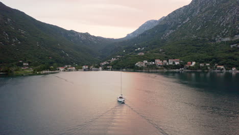 A-luxurious-white-yacht-with-a-tall-mast-without-sails-towing-an-empty-motorboat-and-heading-for-the-port-of-a-small-coastal-town-in-Kotor-bay-in-Montenegro,red-glow-on-the-water-and-mountains-around