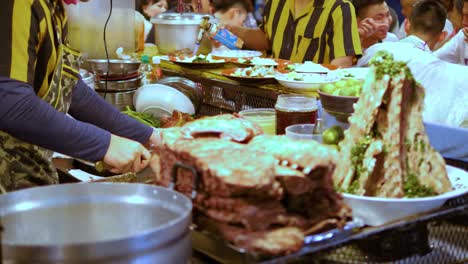 Hombre-De-Uniforme-Negro-Y-Amarillo-Cortó-La-Carne-En-Un-Concurrido-Puesto-Nocturno-En-El-Mercado-De-Alimentos,-Bangkok