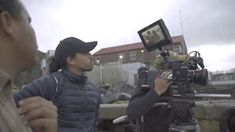 Handgehaltene-Mittlere-Aufnahme-Eines-Filmteams-Mit-Kamera-Und-Crew-Beim-Filmen-Auf-Einem-Holzsteg-In-Hout-Bay-In-Südafrika-Vor-Einem-Wolkigen,-Dunstigen-Hafenpanorama-Mit-Häusern-Und-Bergen-Im-Hintergrund