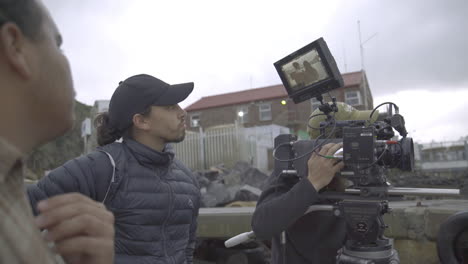 hand-held-medium-shot-of-a-film-crew-with-camera-and-crew-filming-on-a-wooden-pier-in-Hout-Bay-South-Africa-against-a-cloudy-hazy-harbor-panorama-with-houses-and-mountains-in-background