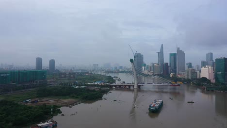 Drohnenaufnahme-Der-Thu-Thiem-2-Brücke-Ist-Eine-Im-Bau-Befindliche-Brücke-In-Ho-Chi-Minh-Stadt-Mit-Einem-Boot,-Das-Schiffscontainer-Transportiert,-Und-Der-Skyline-Der-Stadt