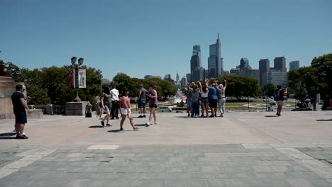 Group-Of-People-Taking-Photos-At-The-Rocky-Steps-In-Pennsylvania---Washington-Monument-Fountain-And-Philadelphia-Skyline-In-Background---timelapse,-wide-shot