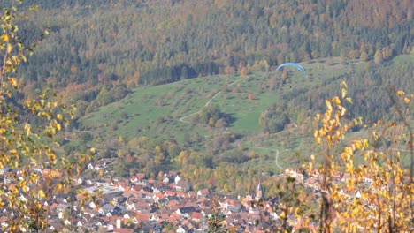 Parapente-Sobrevolando-Una-Pequeña-Ciudad-Y-Un-Impresionante-Paisaje-Otoñal-En-El-Teufelsmühle-En-La-Selva-Negra,-Alemania