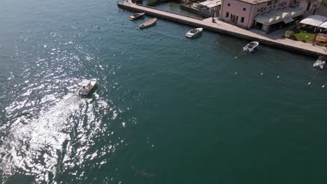 Aerial-view-of-boat-sailing-on-lake-Garda-by-Sato-city-promenade,-Lombardy-Italy-on-sunny-summer-day,-tilt-down-drone-shot