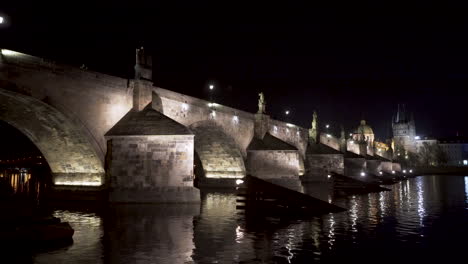 A-view-of-the-stone-arches-of-Charles-bridge-over-river-Vltava-in-the-historical-centre-of-Prague,-Czechia,-at-night,-lit-by-street-lanterns,-reflected-on-the-shimmering-river-surface,-pan-4k-shot