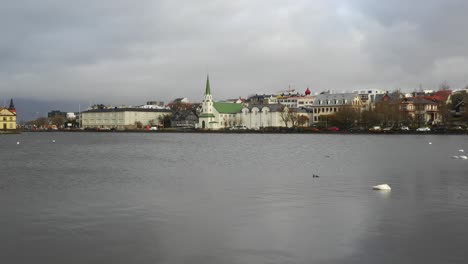 Volando-Sobre-El-Lago-Tjonin-Por-La-Iglesia-Luterana-Frikrikjan-En-La-Ciudad-De-Reykjavik-Islandia-En-Un-Día-Nublado-De-Verano,-Disparo-De-Drones